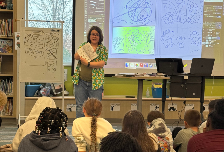 Lindsay Hornsby in a school library giving a comics presentation to 4th/5th graders. She's holding up a comic and pointing to specific examples as the projector displays some of her work-in-progress comics pages from Clip Studio Pro.